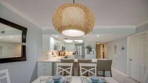 A dining area with a table set for two, a hanging light fixture, and a kitchen in the background. - Pelican Watch #205 Stunning Beachfront Condo (Fort Myers Beach)