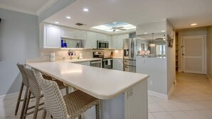 A modern kitchen with white cabinetry, a central island, and a dining area with wicker chairs. - Pelican Watch #205 Stunning Beachfront Condo (Fort Myers Beach)