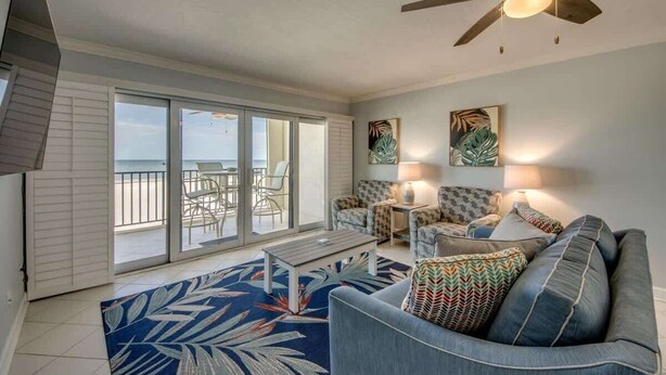 A living room with a sofa, a coffee table, and a ceiling fan. There is a sliding glass door leading to a balcony with a view of the ocean. - Pelican Watch #205 Stunning Beachfront Condo (Fort Myers Beach)