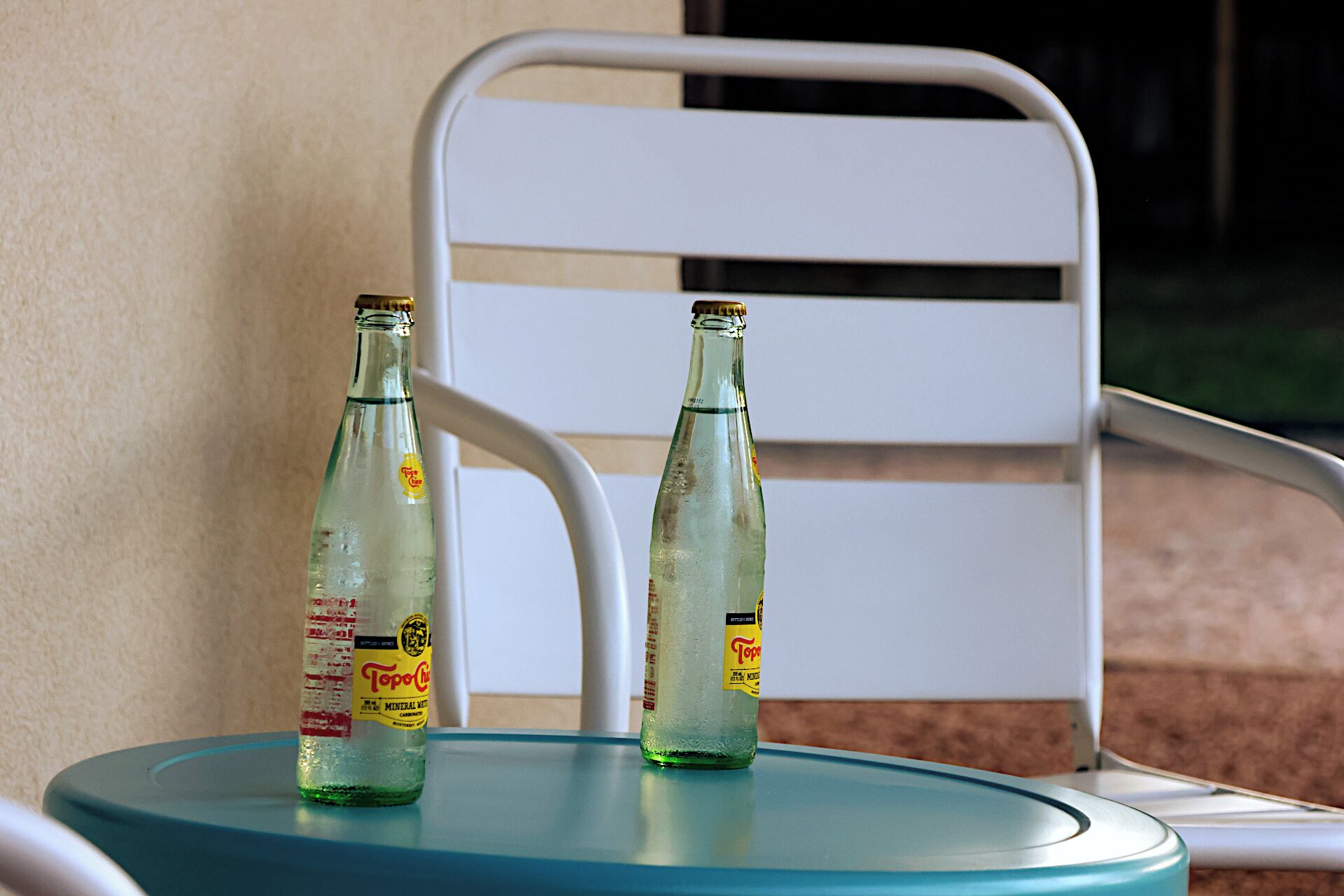 Deux bouteilles en verre vides sont posées sur une table de couleur turquoise, avec une chaise blanche en arrière-plan.
