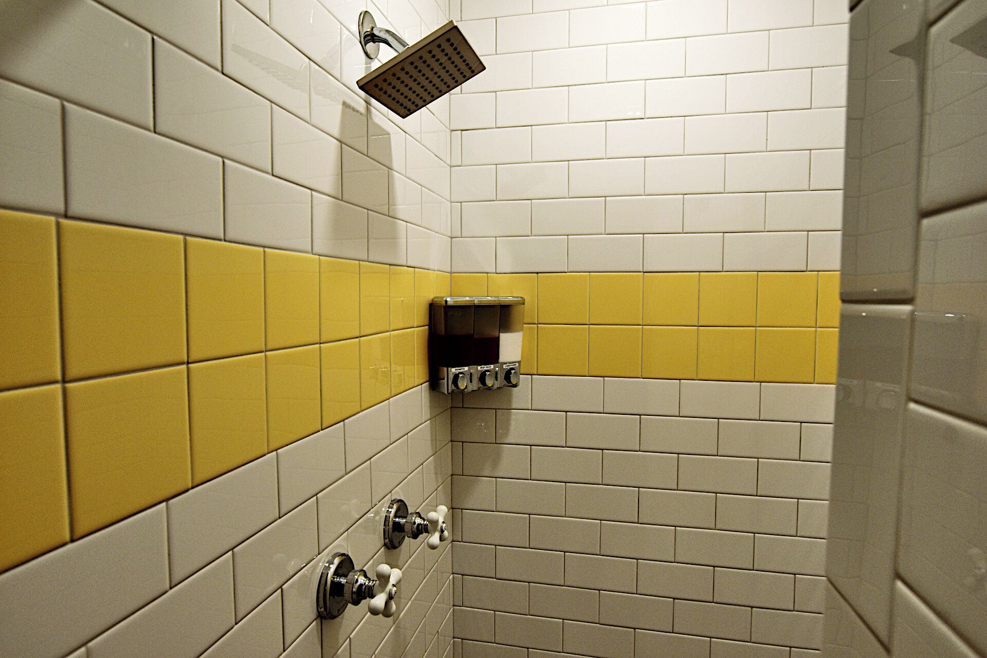 Un baño con azulejos blancos y amarillos, un cabezal de ducha y un perchero para toallas.