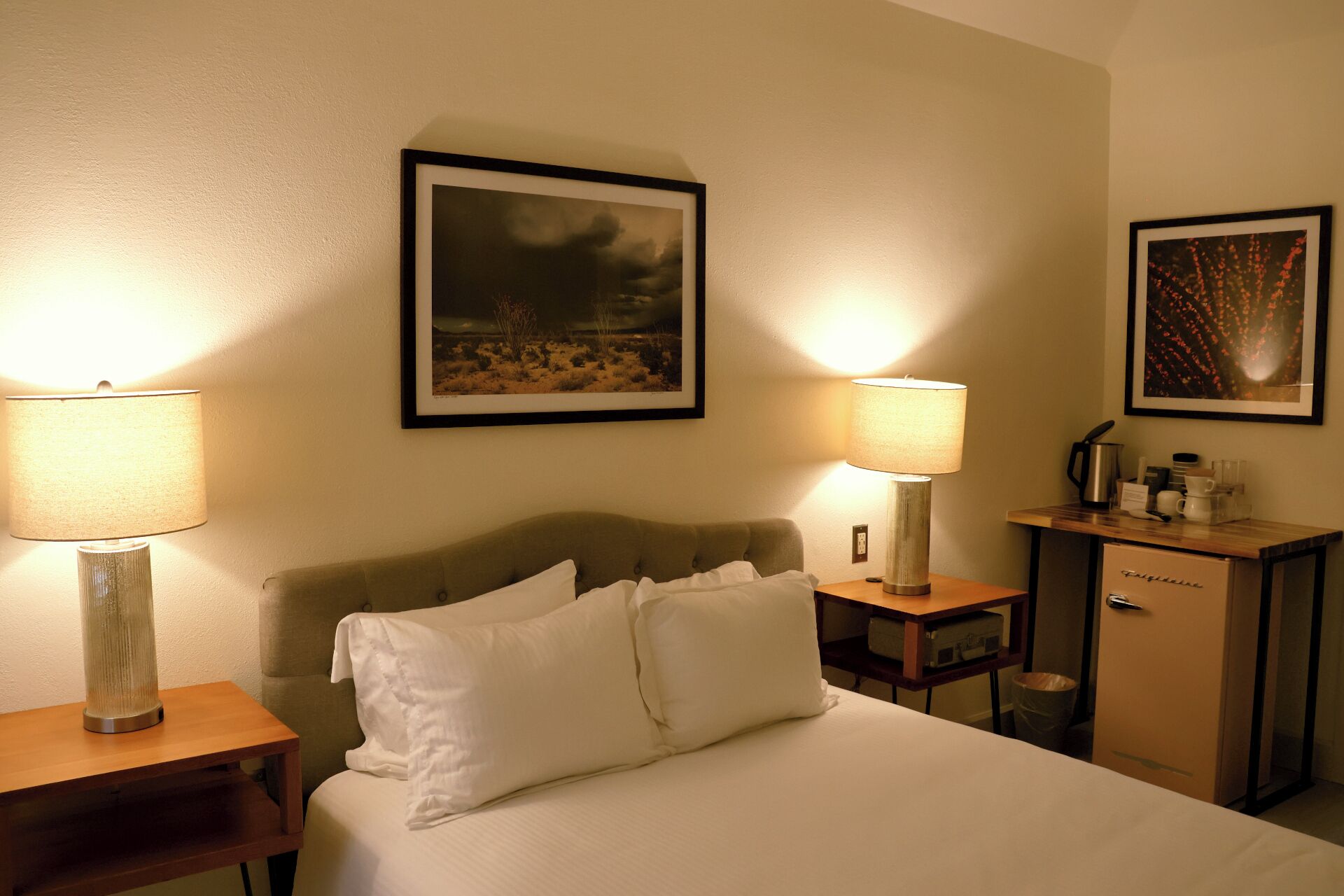 A hotel room with a bed, two bedside lamps, a framed landscape painting, a minibar, and a small table with a lamp.