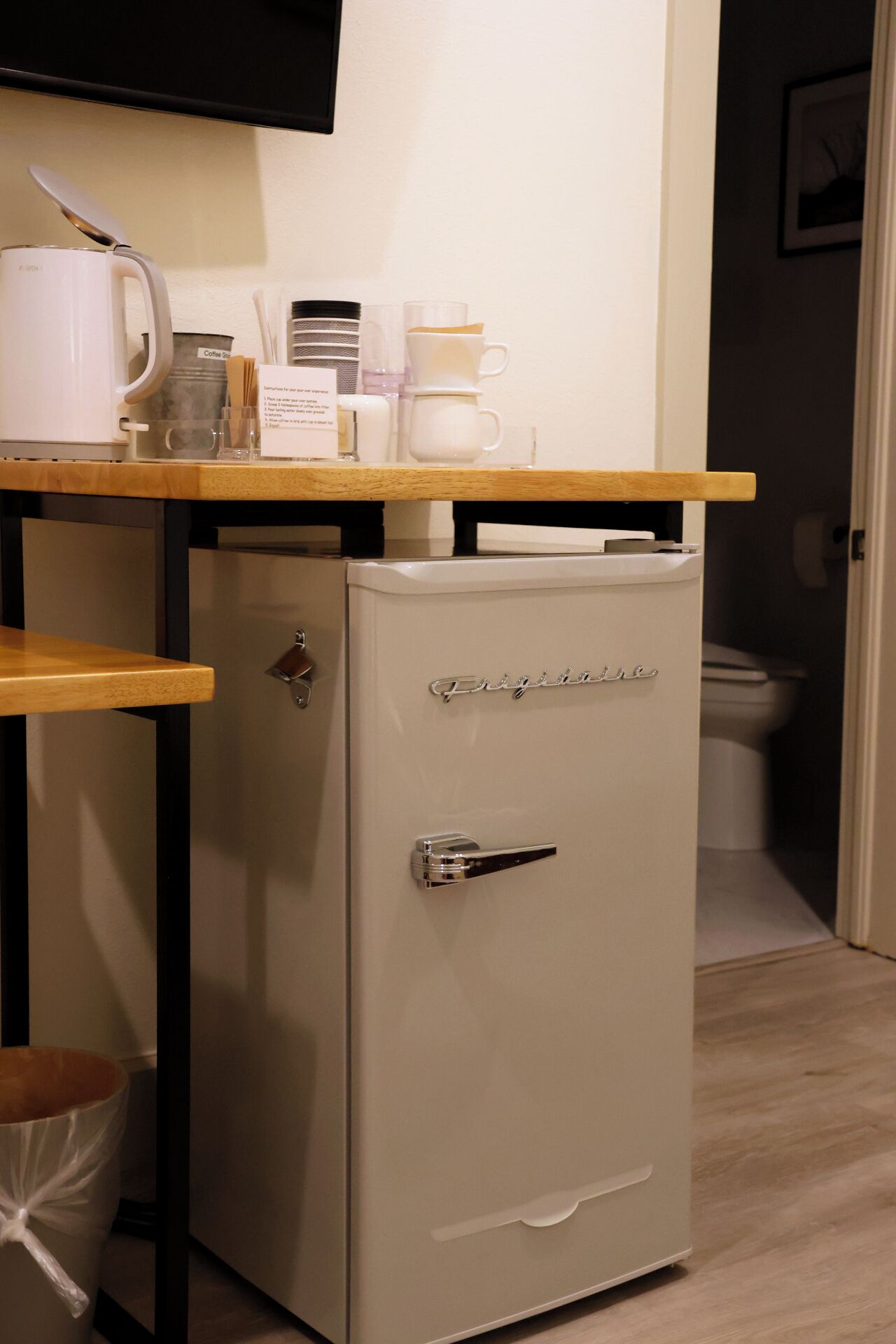 A compact refrigerator with a handle and a nameplate, a coffee maker, a cup, and a small trash bin.