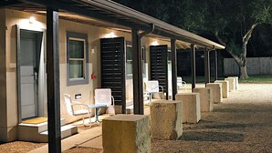 A covered outdoor seating area with benches and tables, a building with a door and windows, and a tree in the background. - The Dixie Motel Brenham (Brenham)