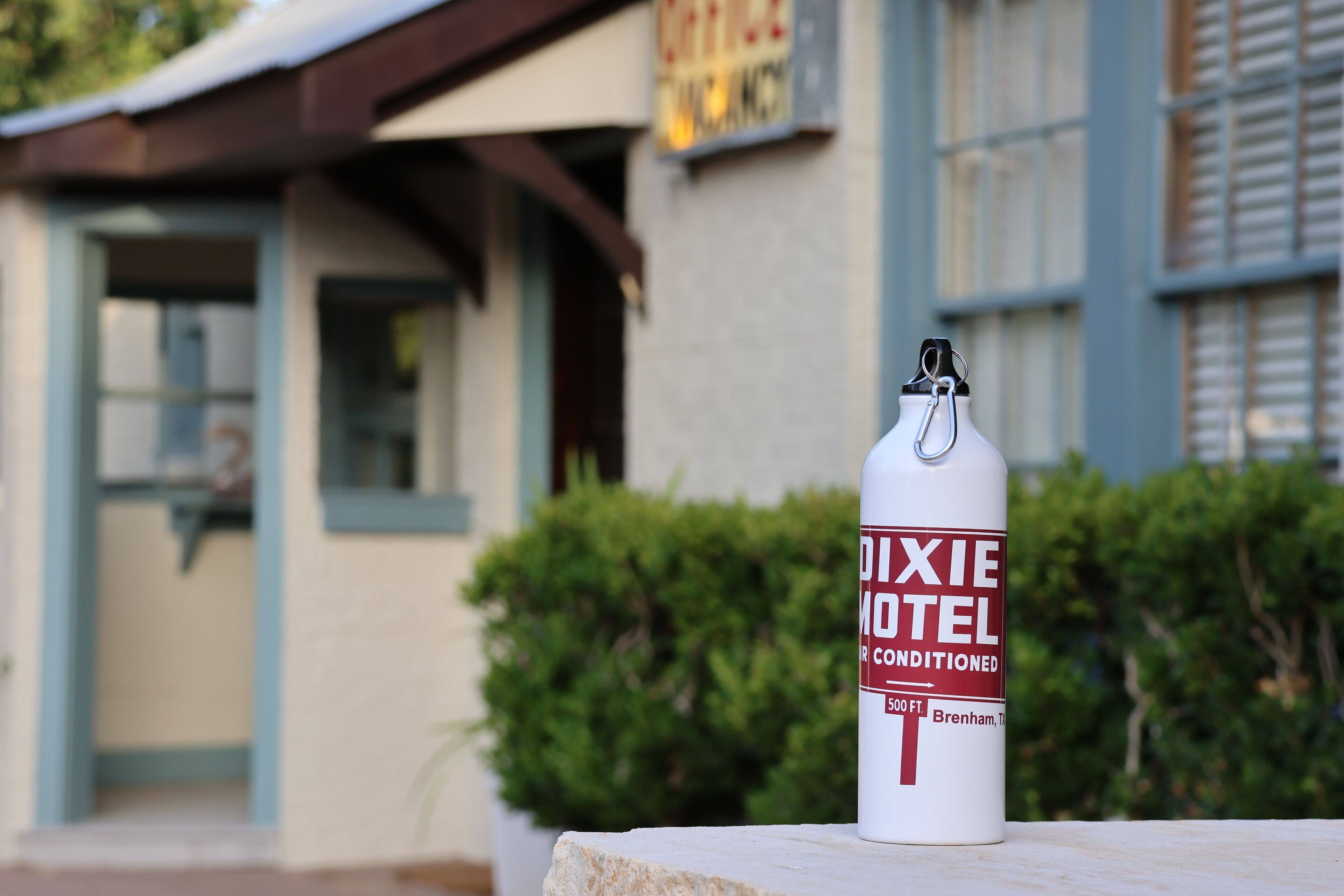 Una botella de agua con una etiqueta que dice "DIXIE HOTEL" y "50 FT.", frente a un edificio con un letrero que indica "DIXIE HOTEL".