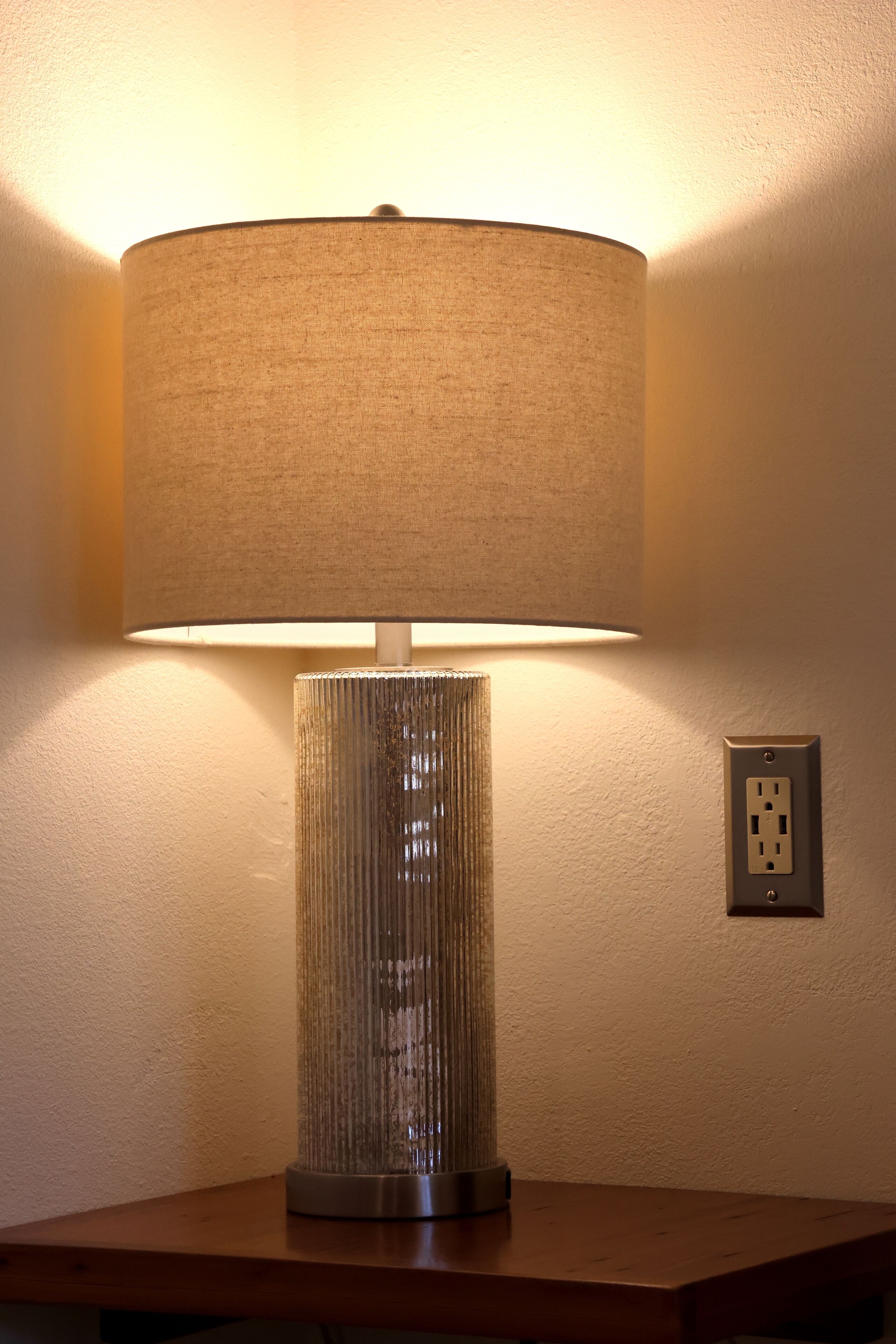 A lamp with a textured lampshade on a wooden table against a plain wall with a power outlet.