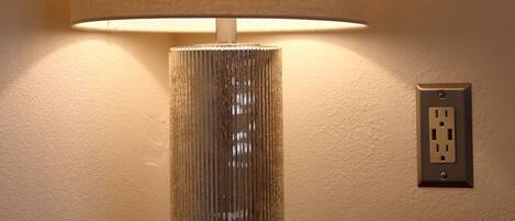 A lamp with a textured lampshade on a wooden table against a plain wall with a power outlet.