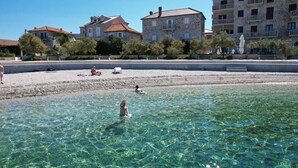 Een strand met helder water, mensen die zwemmen en spelen, en gebouwen op de achtergrond.
