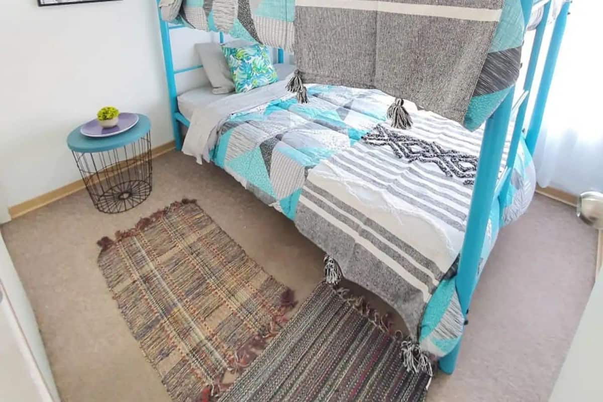 A bedroom with a blue metal bed frame, a patterned bedspread, two pillows, a small round table with a plant, and a cityscape artwork on the wall.