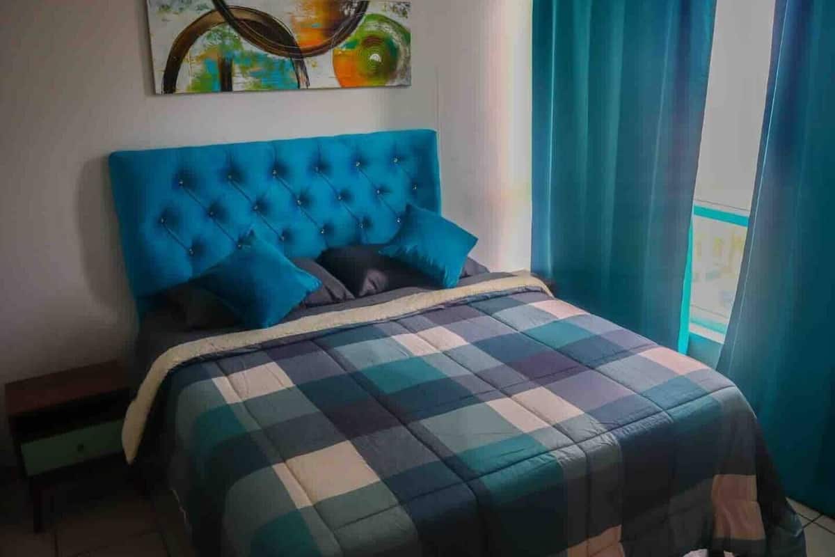 A bedroom with a blue tufted headboard, a plaid bedspread, and a painting above the bed.