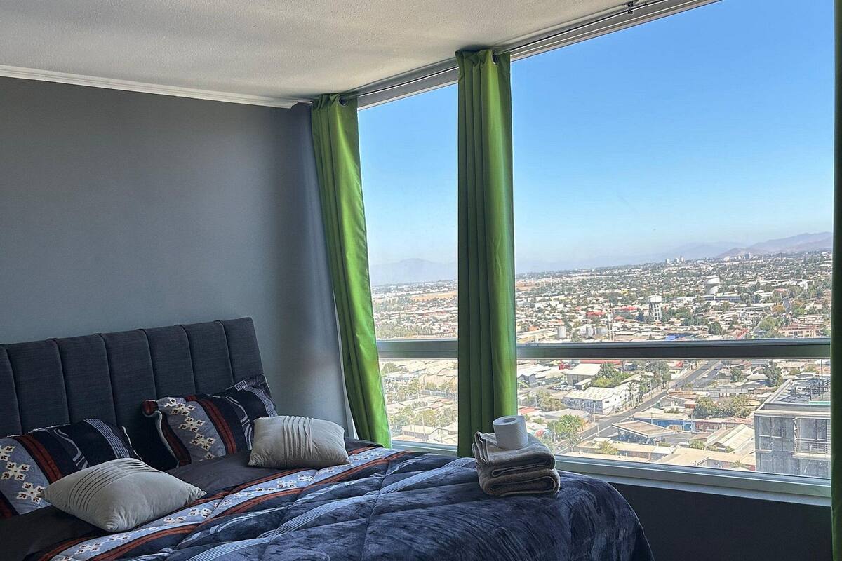 A bedroom with a large bed, green curtains, and a city view.