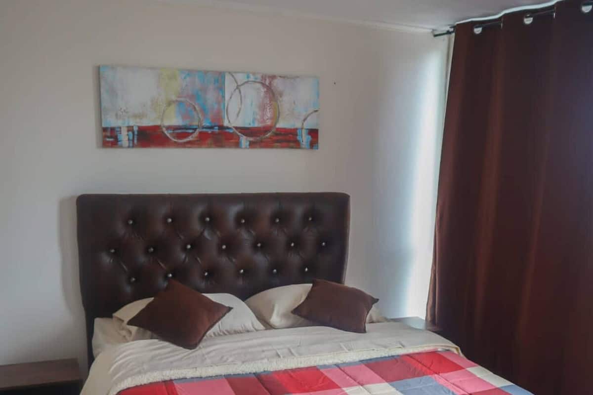 A bedroom with a large bed, a headboard, a nightstand, a painting on the wall, and a window with curtains.