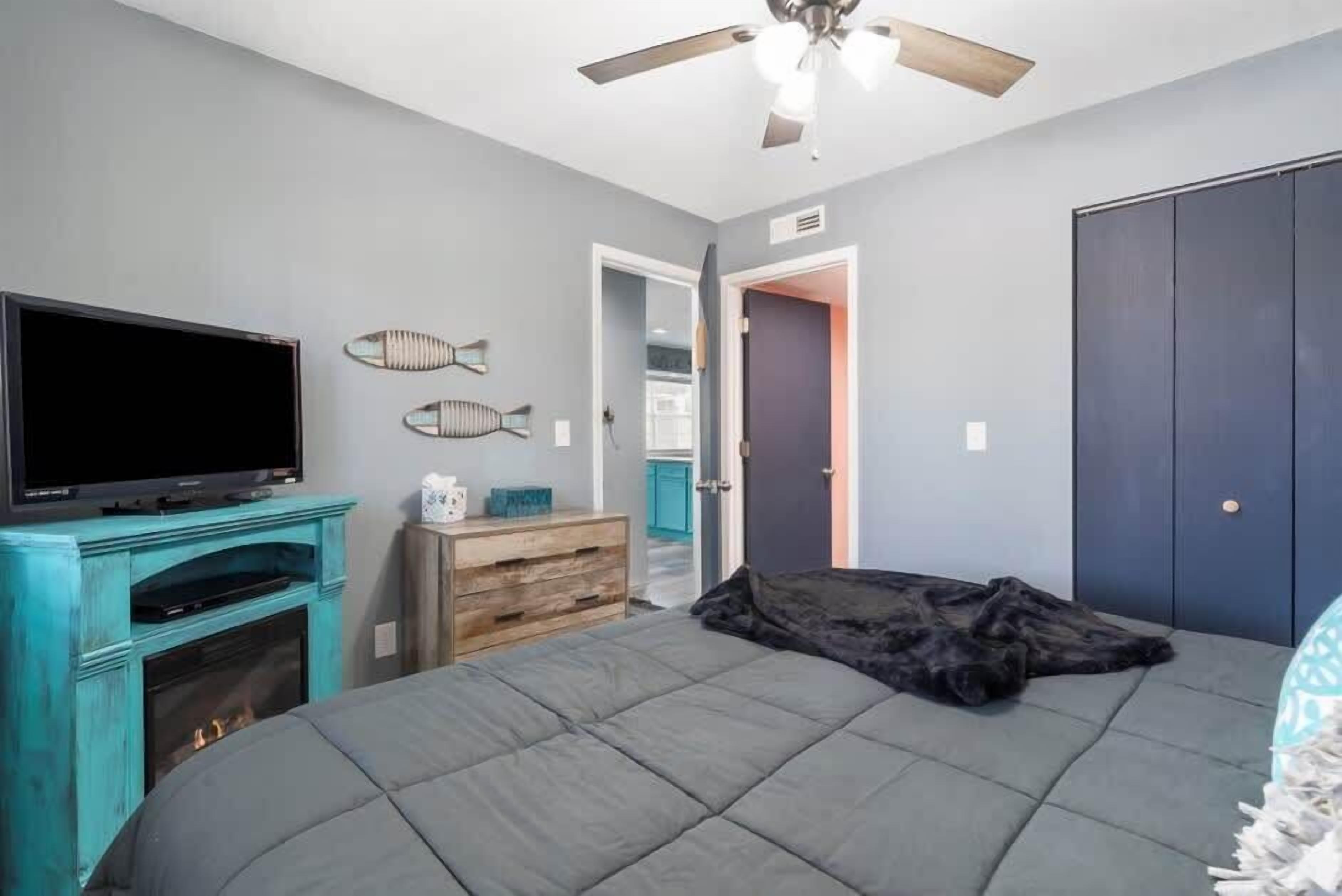 A bedroom with a bed, a TV, a ceiling fan, and a door leading to another room.
