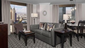 Una habitación de hotel moderna con un sofá gris, una mesita auxiliar pequeña y un área de comedor con una mesa y sillas. Se aprecia una vista panorámica de la ciudad a través del amplio ventanal.