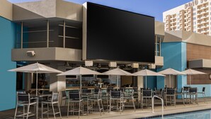 A poolside bar area with outdoor seating and umbrellas, a large flat-screen TV, and a bar with a variety of bottles.