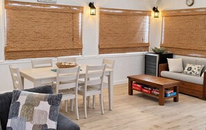 A room with a dining table, chairs, a sofa, and a coffee table. There are two windows with blinds, a wall clock, and a wall-mounted lamp.