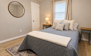 A bedroom with a bed, a bedside table, a lamp, a window with curtains, a door, and a mirror on the wall.