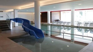 Zona interior de piscina moderna con un tobogán azul, barandillas de cristal y vistas a una zona de estar exterior.