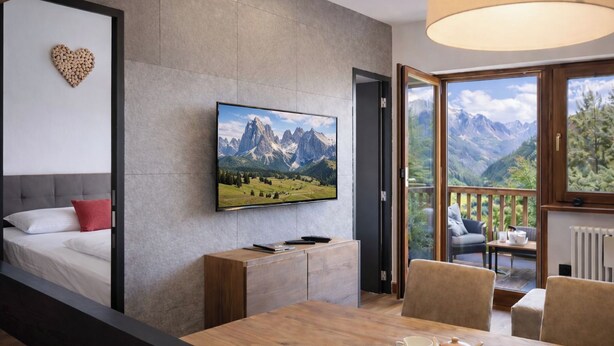 Una habitación de hotel moderna con cama, televisor montado en la pared, una mesa de comedor con sillas y un balcón con vista a la montaña.