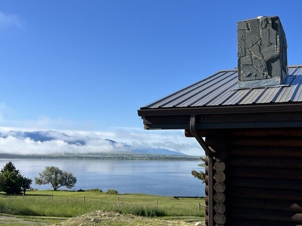 A rustic cabin with a stone chimney, overlooking a calm lake and distant mountains under a clear sky.