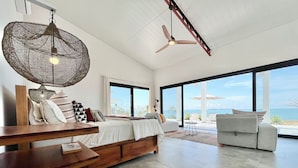 Une chambre à coucher moderne avec un grand lit, un banc en bois, un ventilateur de plafond et une vue sur la mer.