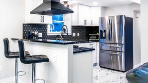 A modern kitchen with white cabinets, a stainless steel refrigerator, and black bar stools. - Gatsby Theme Near Downtown w Game Room, EV Charger (Lake Geneva)
