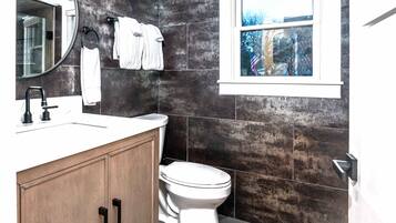 A bathroom with a white sink, dark tiled walls, a toilet, and a window with a view of trees and an American flag.