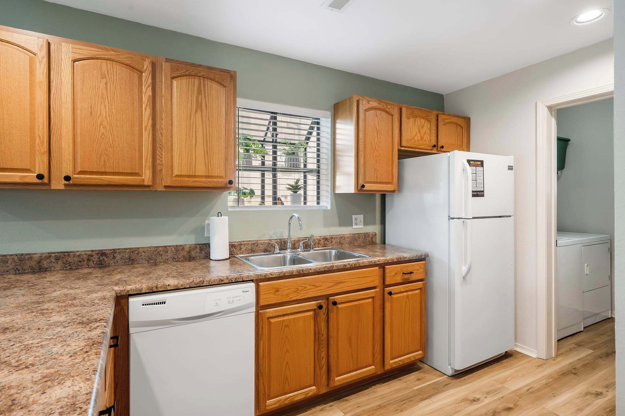 A kitchen with wooden cabinets, a white refrigerator, a dishwasher, and a window with blinds.