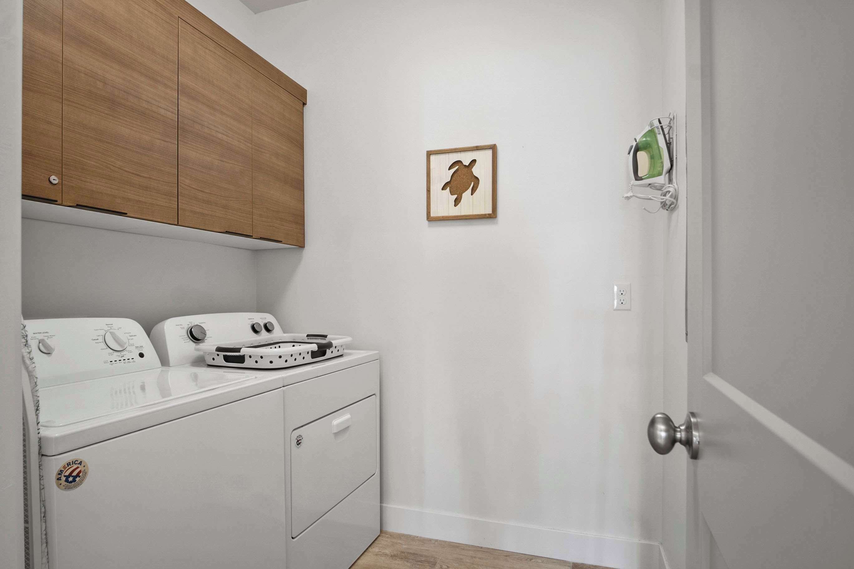A laundry room with a front-loading washer and dryer, wooden cabinets above, a framed turtle picture on the wall, and a green soap dispenser on the door.