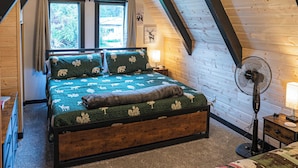 Un dormitorio acogedor en el ático con techo inclinado, una cama con colcha verde estampada, cabecera de madera, un ventilador de pie y una ventana con cortinas.