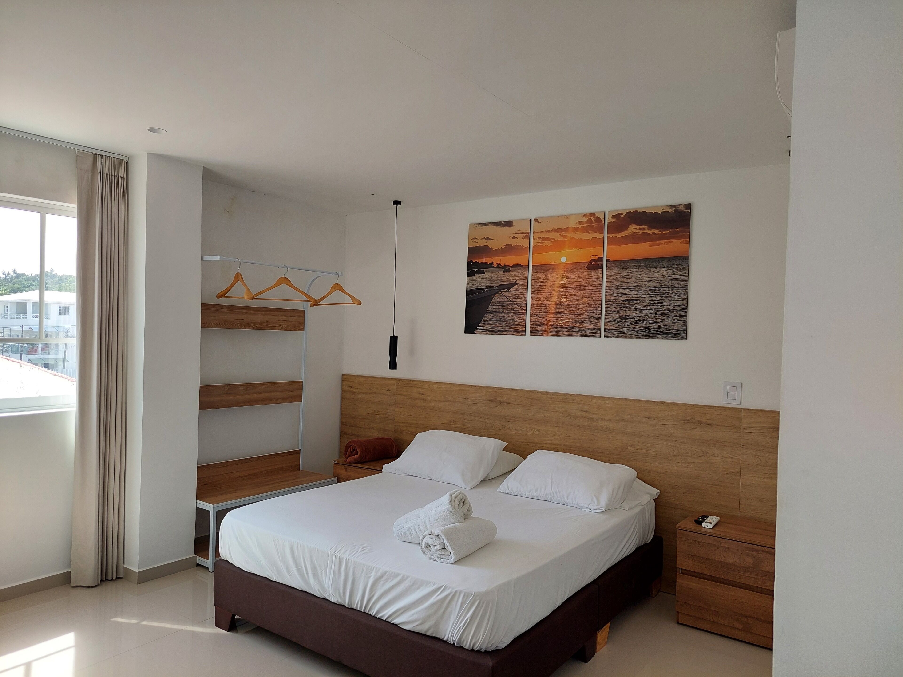 Un dormitorio moderno con cabecera de madera, cama con ropa blanca, mesita de noche, cómoda de madera, armario con estantes de madera y un ventanal con cortinas.