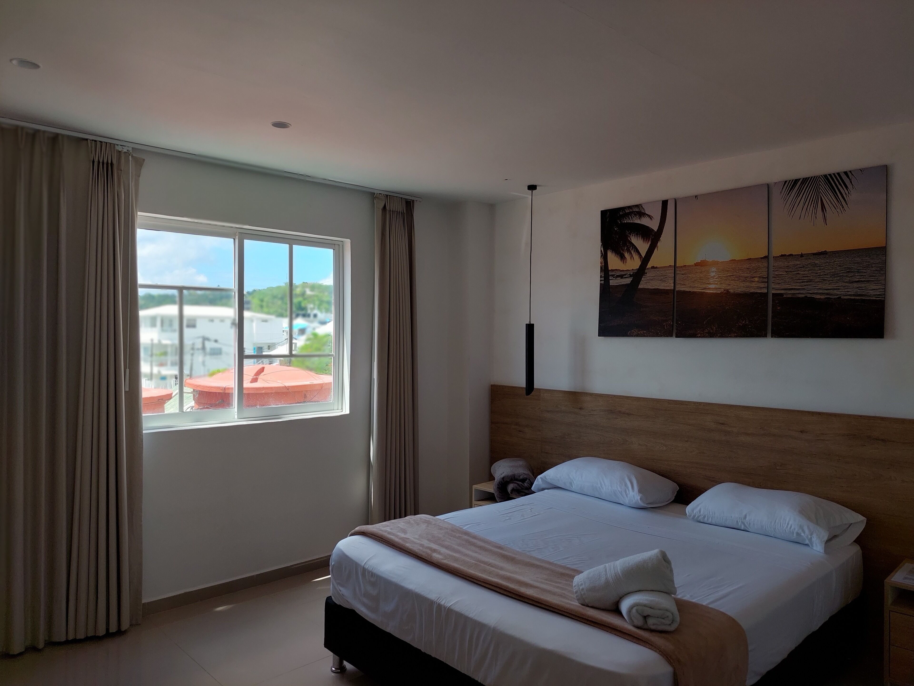 Hotellihuone, jossa on sänky, yöpöytä, ikkuna verhoilla ja taulu, jossa on kuvattuna auringonlasku meren yllä.