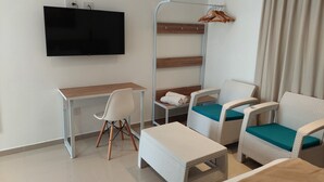 Una habitación de hotel moderna con televisión de pantalla plana, un escritorio, dos sillas de mimbre con cojines color aguamarina, una mesita blanca y un estante de madera.