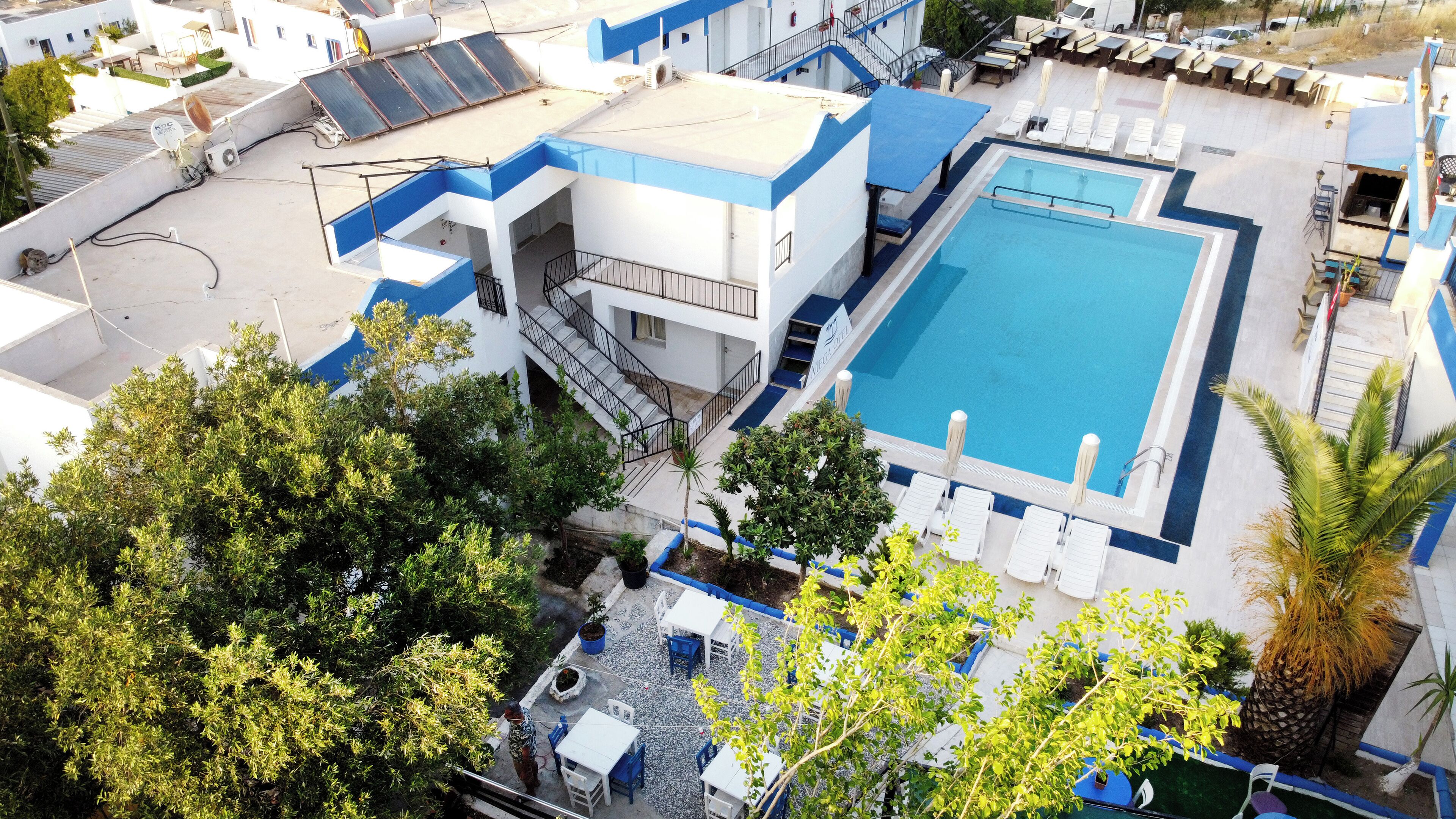 Un área de piscina en la azotea con un edificio azul y blanco, una escalera y follaje circundante.