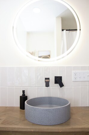 A bathroom with a round sink, a mirror with a light, and a towel rack.