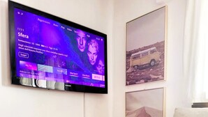 A living room with a flat-screen TV mounted on the wall, a white sofa, a small white table, and a vase with purple flowers. - Close to Franklin College and Lugano (Collina d'Oro)