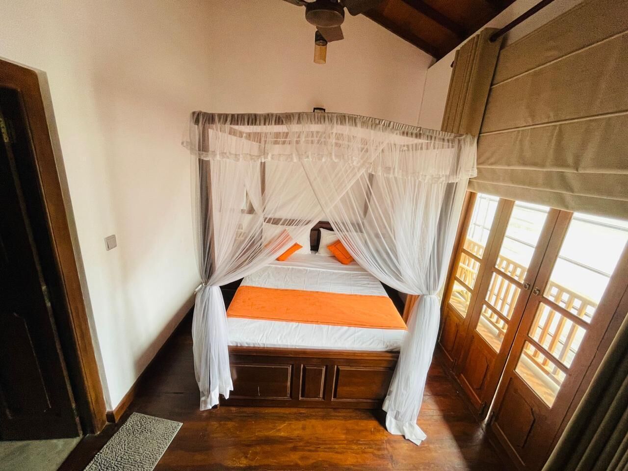 A room with a bed under a canopy, wooden flooring, and a view through a window with a wooden railing.