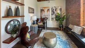 Una sala de estar acogedora con un sillón de cuero, un reposapiés redondo y una mesita de centro. Hay estanterías con adornos y un reloj de pared.