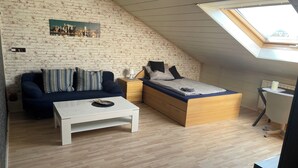 Ein Loft-Zimmer mit schrägem Dach, einem Bett mit Holzbettgestell, einem weißen Couchtisch, einem dunkelblauen Sofa und einem kleinen Schreibtisch mit Lampe.