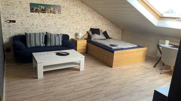 Una habitación estilo loft con techo inclinado, una cama con estructura de madera, una mesa de centro blanca, un sofá azul oscuro y un pequeño escritorio con lámpara.