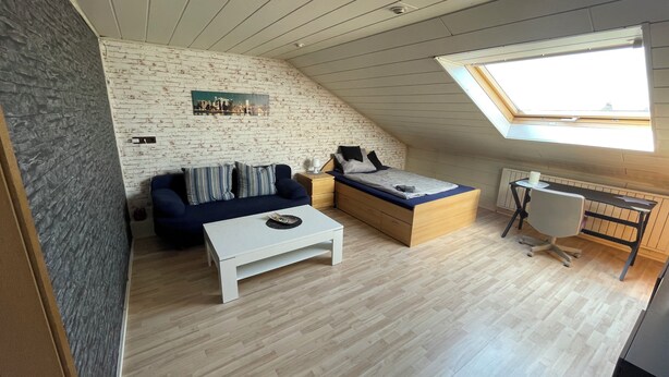 Ein Loft-Zimmer mit schräg verlaufender Decke, einem Bett, einer Couch, einem kleinen Tisch und einem Schreibtisch mit Stuhl.