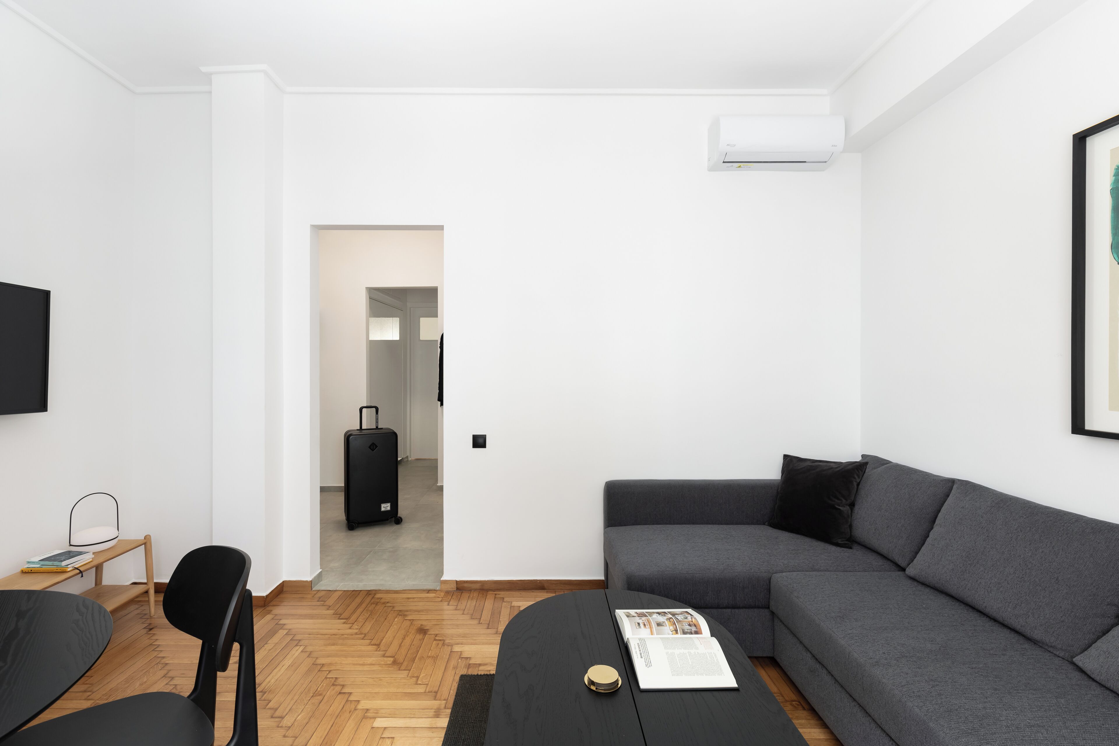 Una sala de estar moderna con un sofá gris, un sillón negro, una mesita con una revista y una maleta negra al fondo.