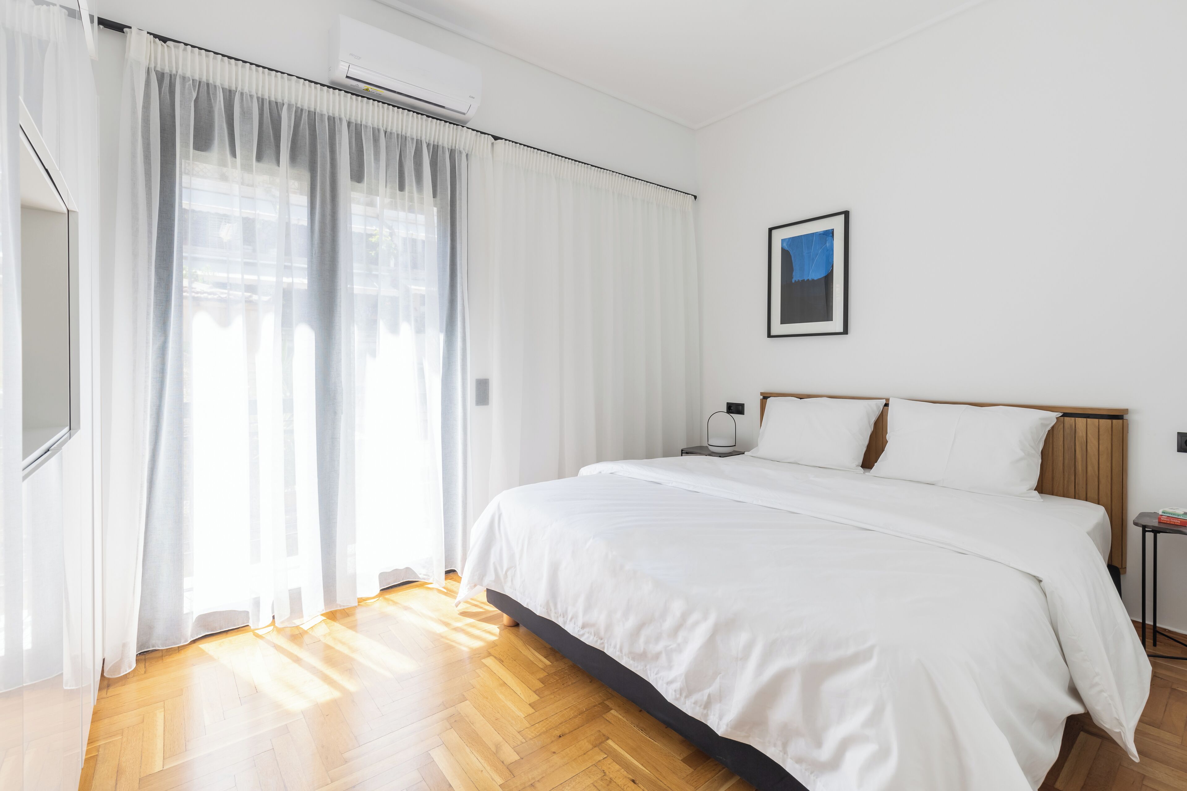 A hotel room with a large bed, white curtains, a framed picture on the wall, and a wooden floor.