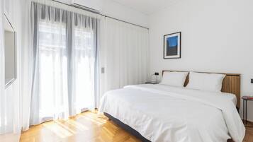 A hotel room with a large bed, white curtains, a framed picture on the wall, and a wooden floor.