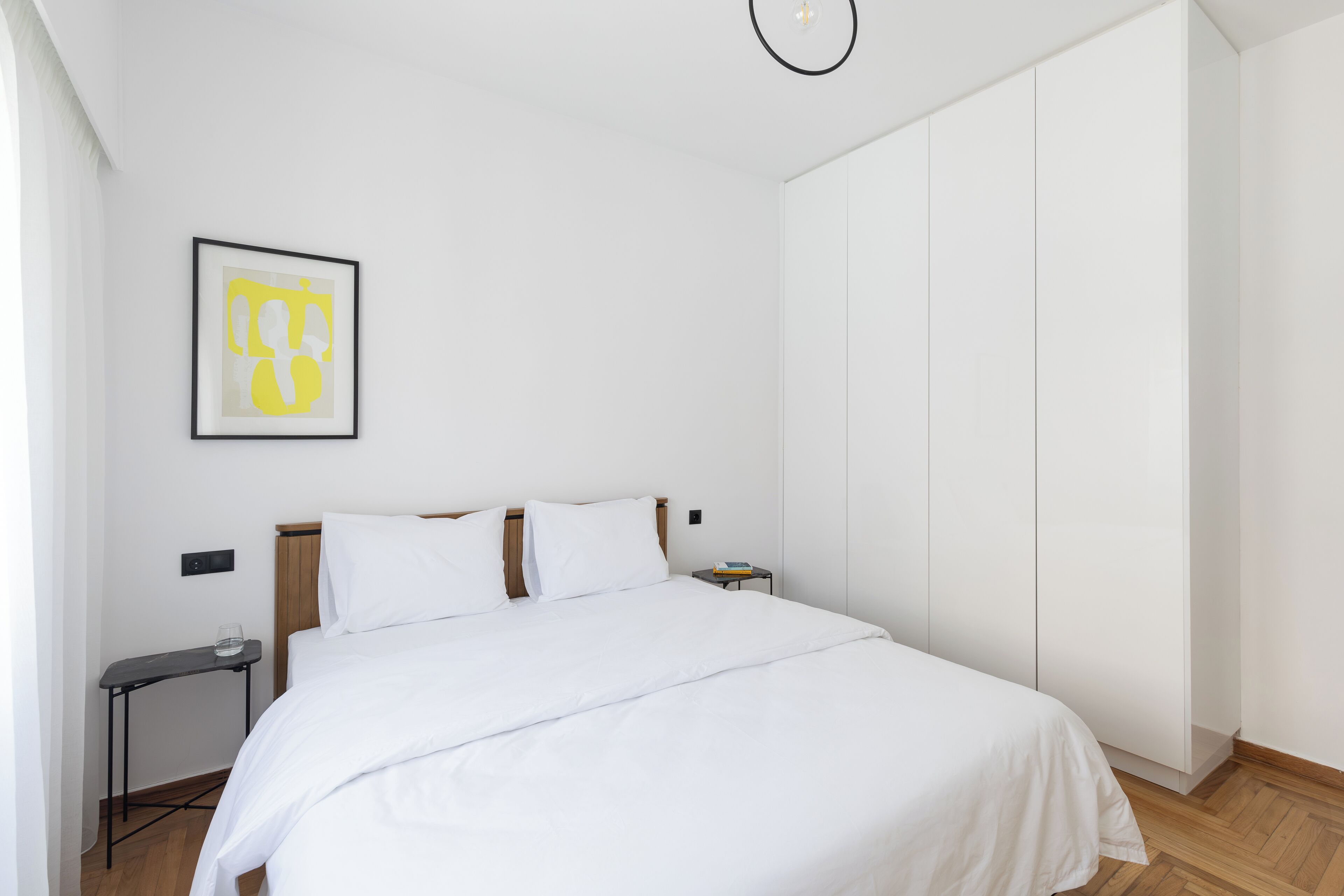 Une chambre à coucher au style minimaliste, avec un grand lit, une petite table de chevet, un tableau encadré au mur et une armoire.