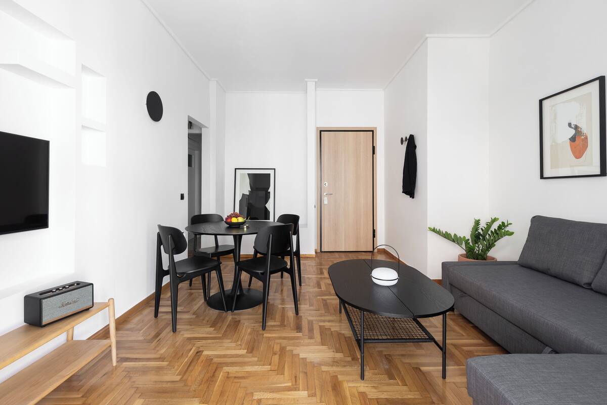 A modern living room with a grey sofa, a dining table with chairs, a TV on the wall, and a wooden floor.