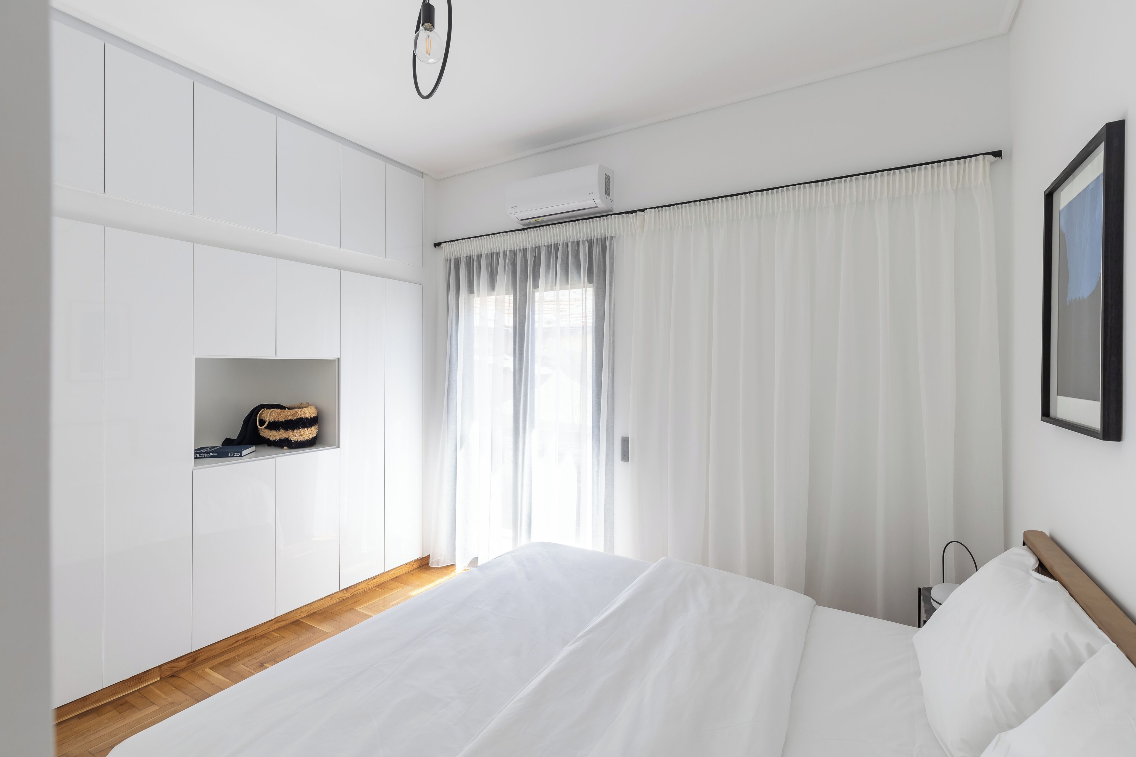 A modern bedroom with a large bed, white walls, a wooden floor, a built-in wardrobe, and a window with sheer curtains.