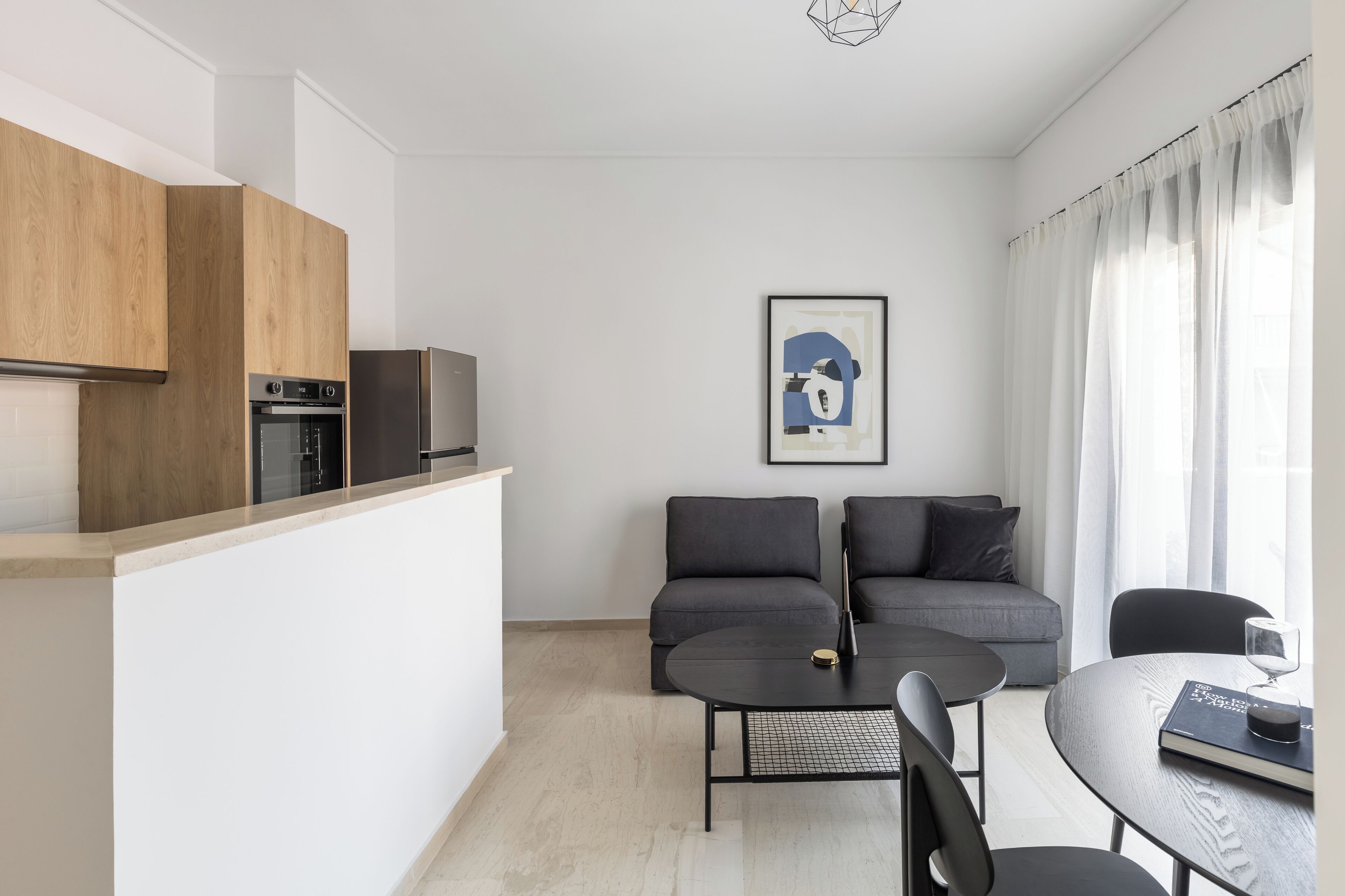 A modern kitchen with wooden cabinets and a built-in oven, a gray sofa set, a dining table with chairs, and a framed artwork on the wall.