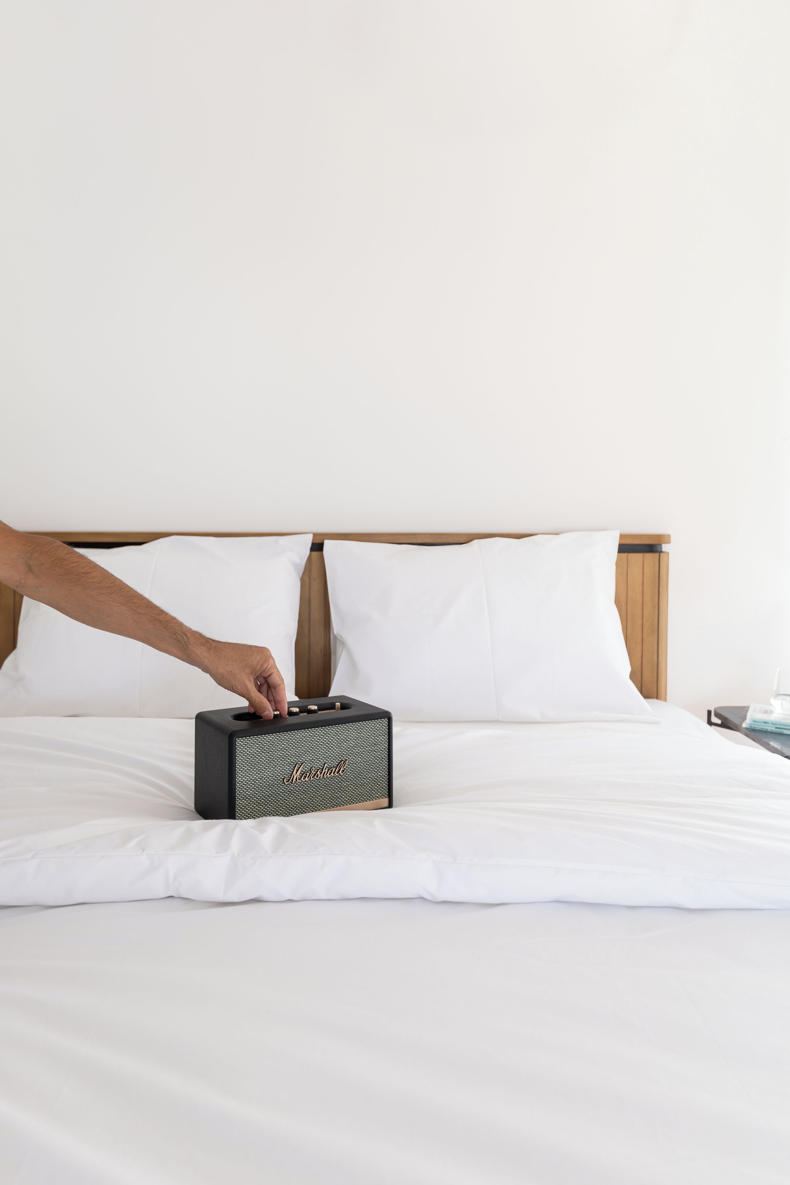 A hand adjusting a portable speaker on a bed with white bedding and a wooden headboard.