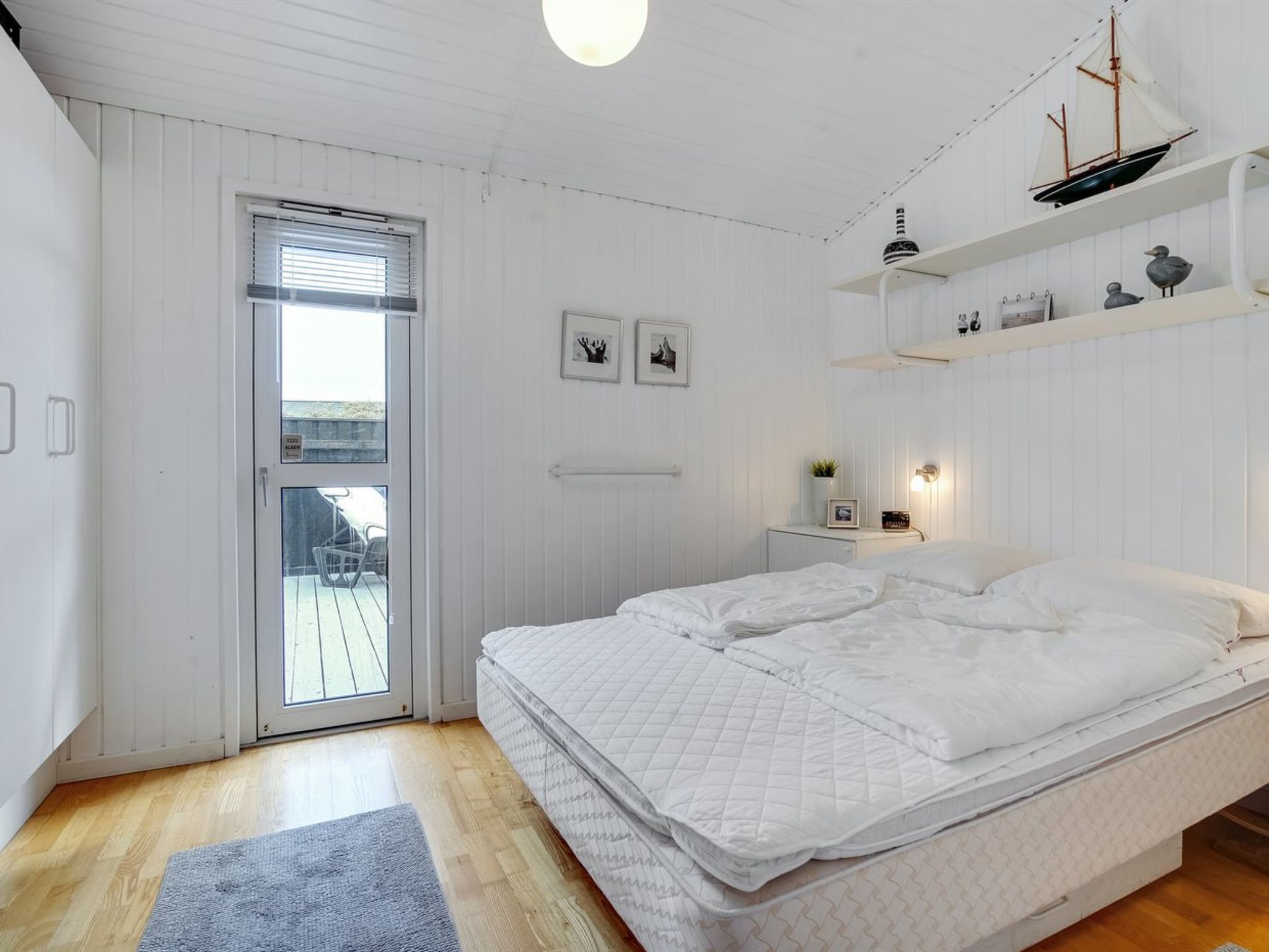 Un dormitorio con cama, una mesita de noche pequeña, un estante con objetos decorativos, una puerta con vistas al exterior y una ventana con persianas.
